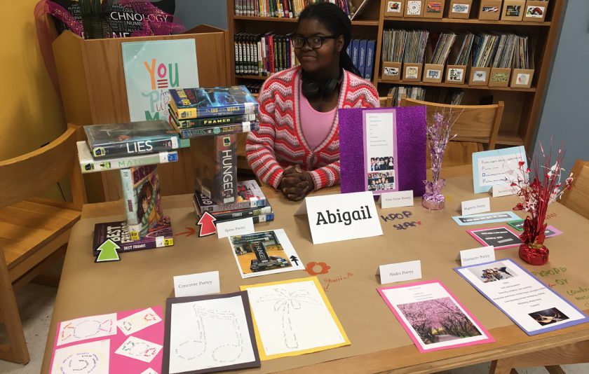 student sits behind a table with her poetry in different colorful forms