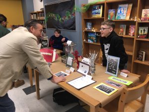 Principal leans in to speak with student behind desk filled with the poems he created