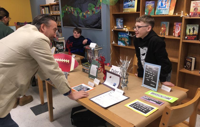 Principal leans in to speak with student behind desk filled with the poems he created