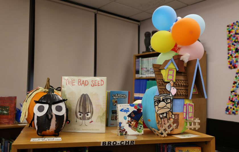 painted pumpkins next to boocks one is from the book Up with a house and an old man and baloons lifting the house and the other pumpkin has big eyes and is black and white striped for the bad seed book