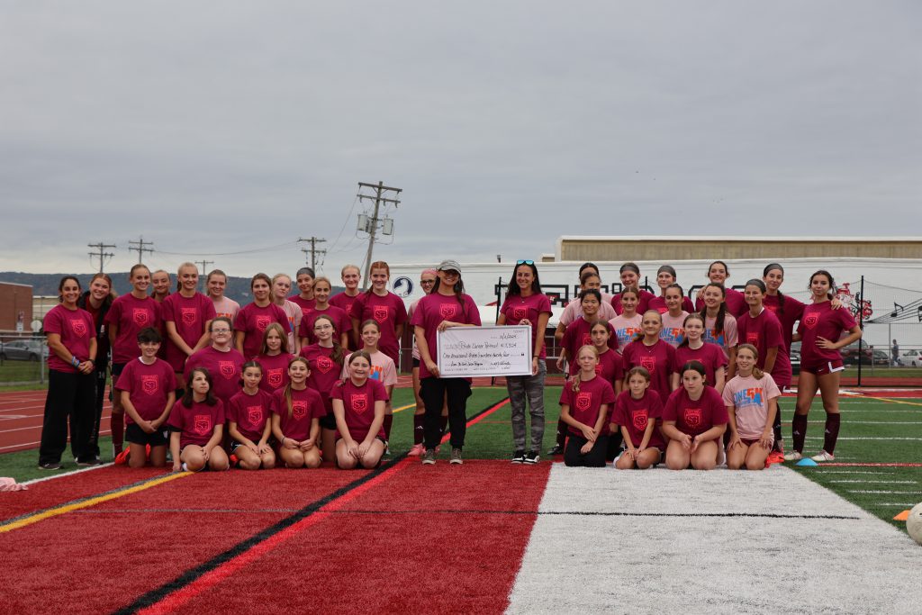 students in red soccer uniforms surrounding coaches and representative accepting a donation in the form of an oversized check