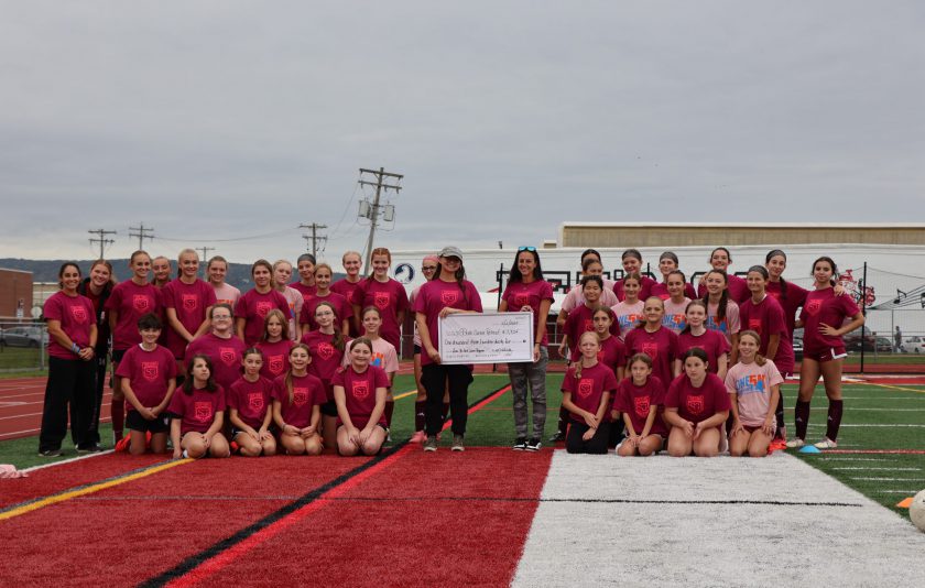 students in red soccer uniforms surrounding coaches and representative accepting a donation in the form of an oversized check