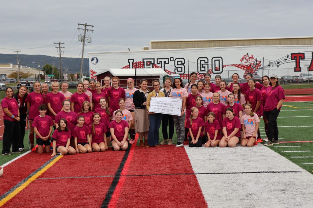 students in red soccer uniforms surrounding coaches and representative accepting a donation in the form of an oversized check