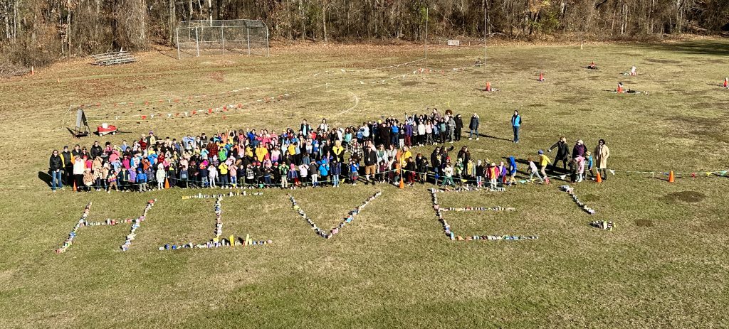 students and teacher and staff standing behind food donations that spell out HIVE on an outdoor field of the school