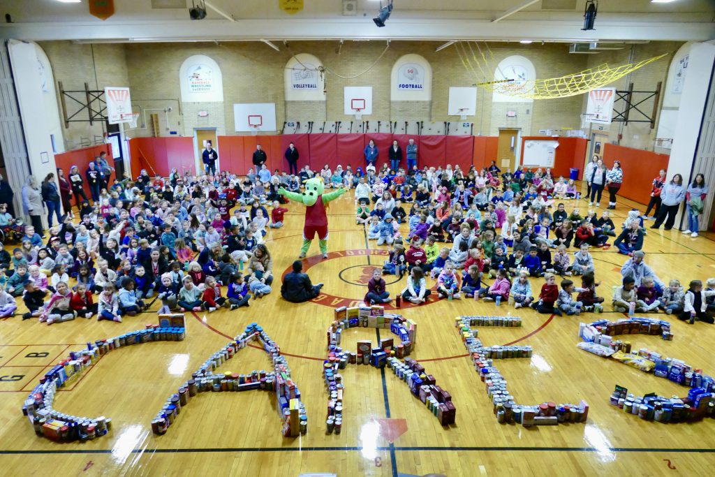 a dragon mascot standing with its hands up in the middle of the gym with students sitting on each side. The food donated is arranged into the word cares. 