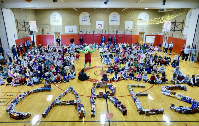 a dragon mascot standing with its hands up in the middle of the gym with students sitting on each side. The food donated is arranged into the word care.