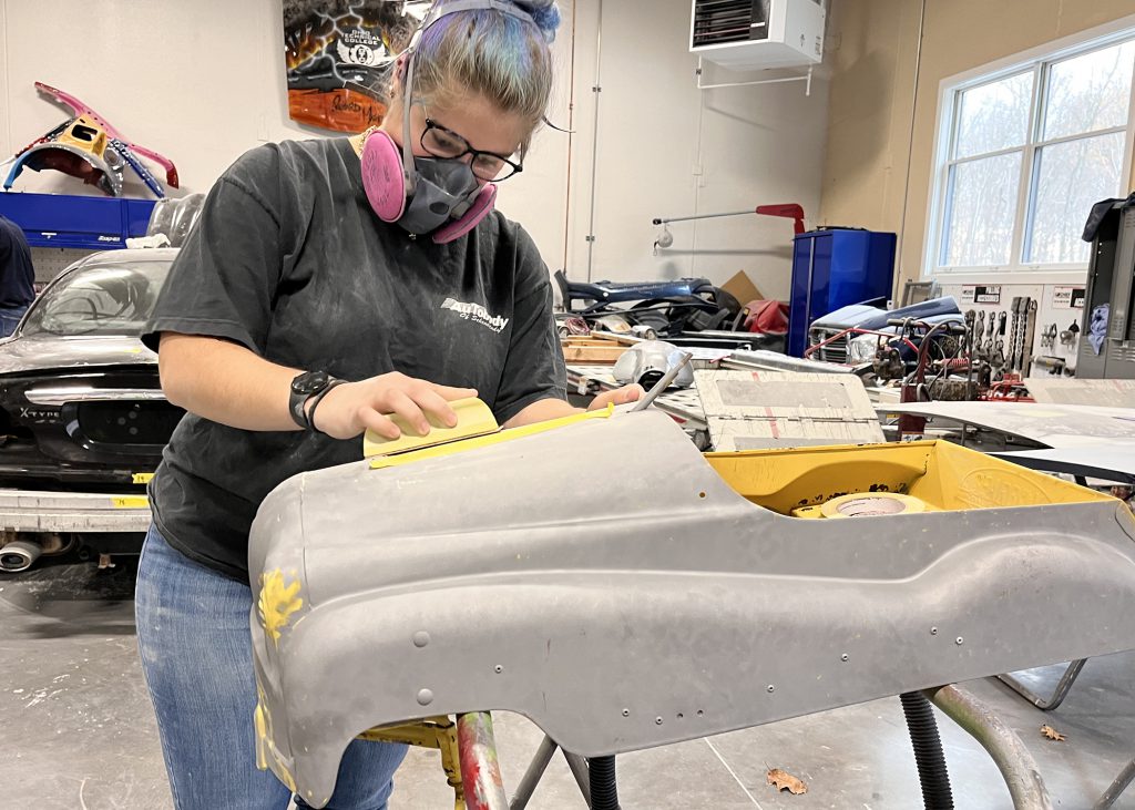 student wearing a protective mask sands down a car part