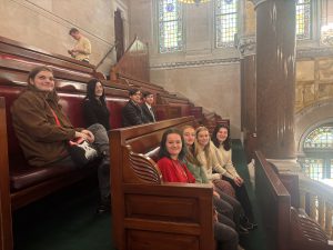 Students looking over at camera to smile while sitting on wood benches in the senate gallery